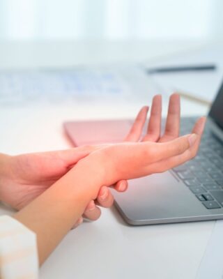 A person holds their wrist in pain whilst sitting at a desk with an open laptop, suggesting discomfort possibly caused by repetitive computer work or carpal tunnel syndrome.