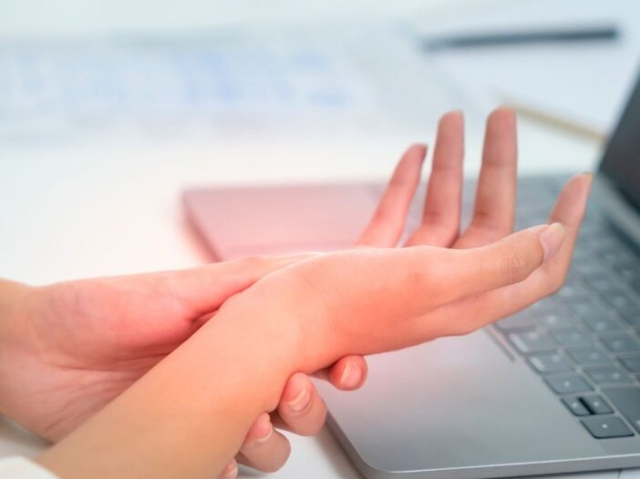 A person holds their wrist in pain whilst sitting at a desk with an open laptop, suggesting discomfort possibly caused by repetitive computer work or carpal tunnel syndrome.
