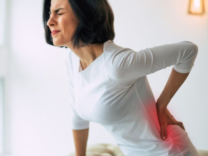 A woman in a white shirt sits on a sofa, holding her lower back and wincing in pain, indicating discomfort likely due to back pain. Warm indoor lighting and a window are in the background, suggesting she may need orthopaedic treatment.